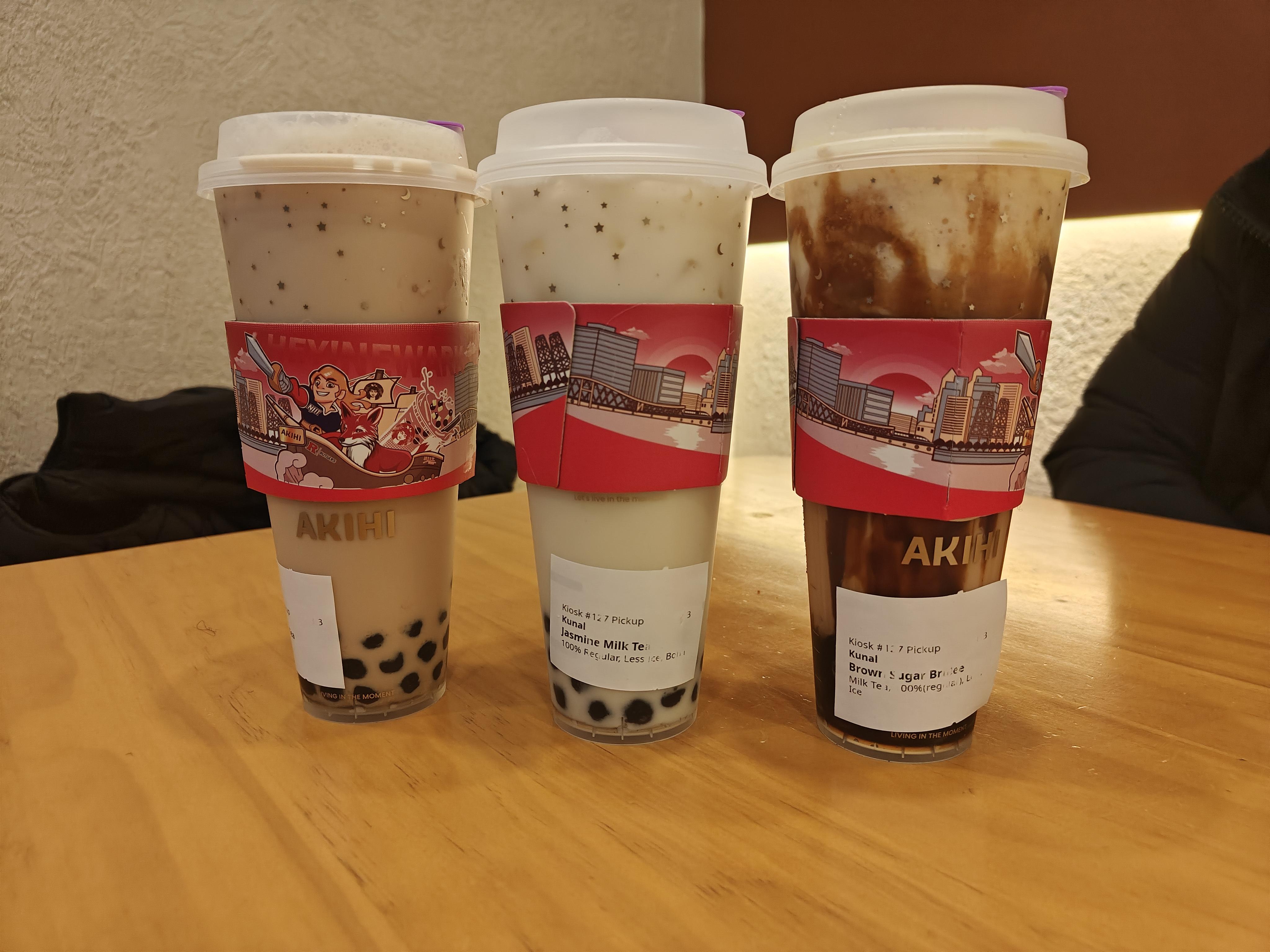 Three cups of boba; with partially readable white labels on the bottom with the drink names. They have red sleeves with a guy holding a sword on a ship and Rutgers in the background