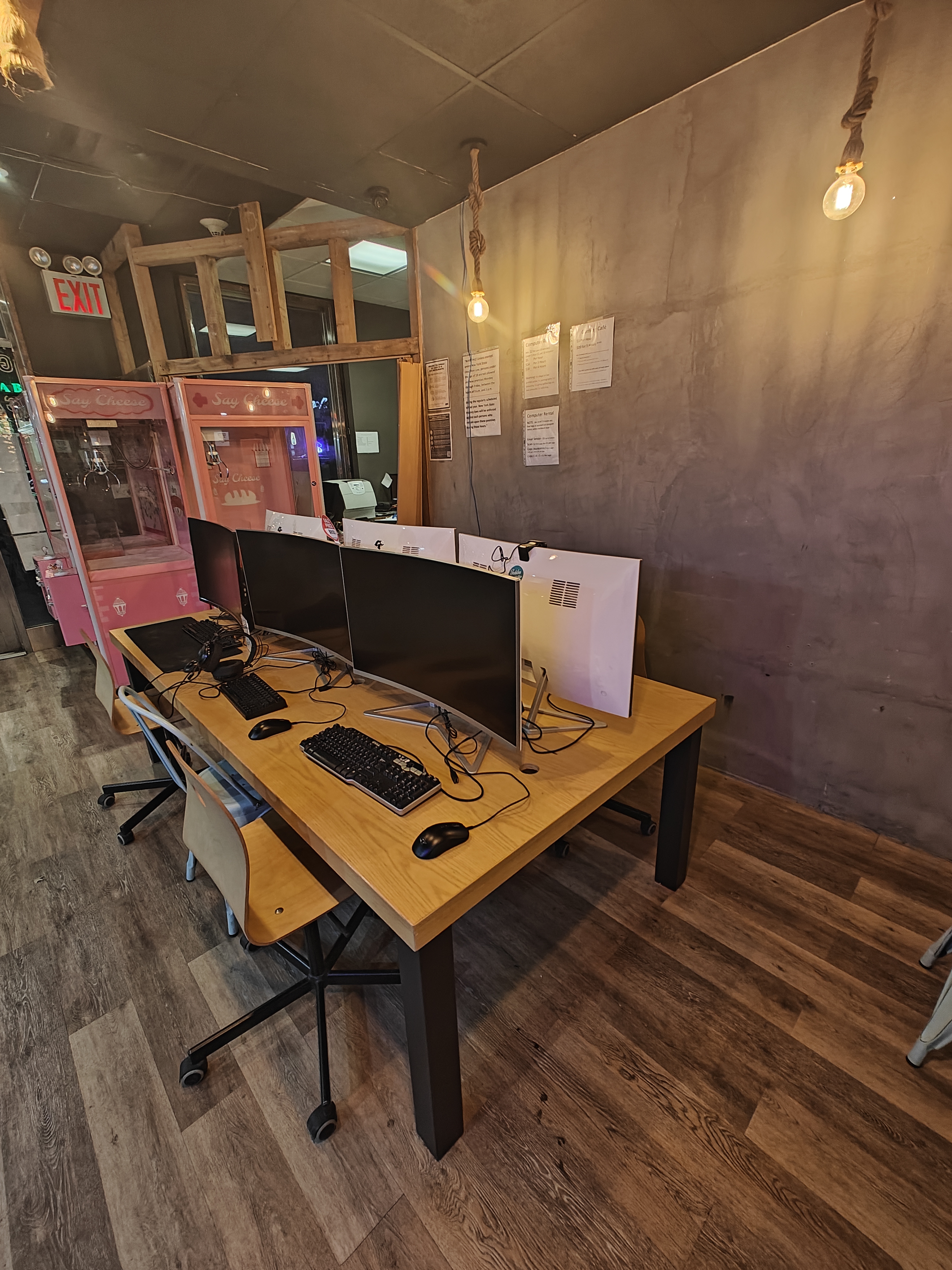 A table with computers on it, in the setup of a gaming cafe. Empty claw machines are visible in the background