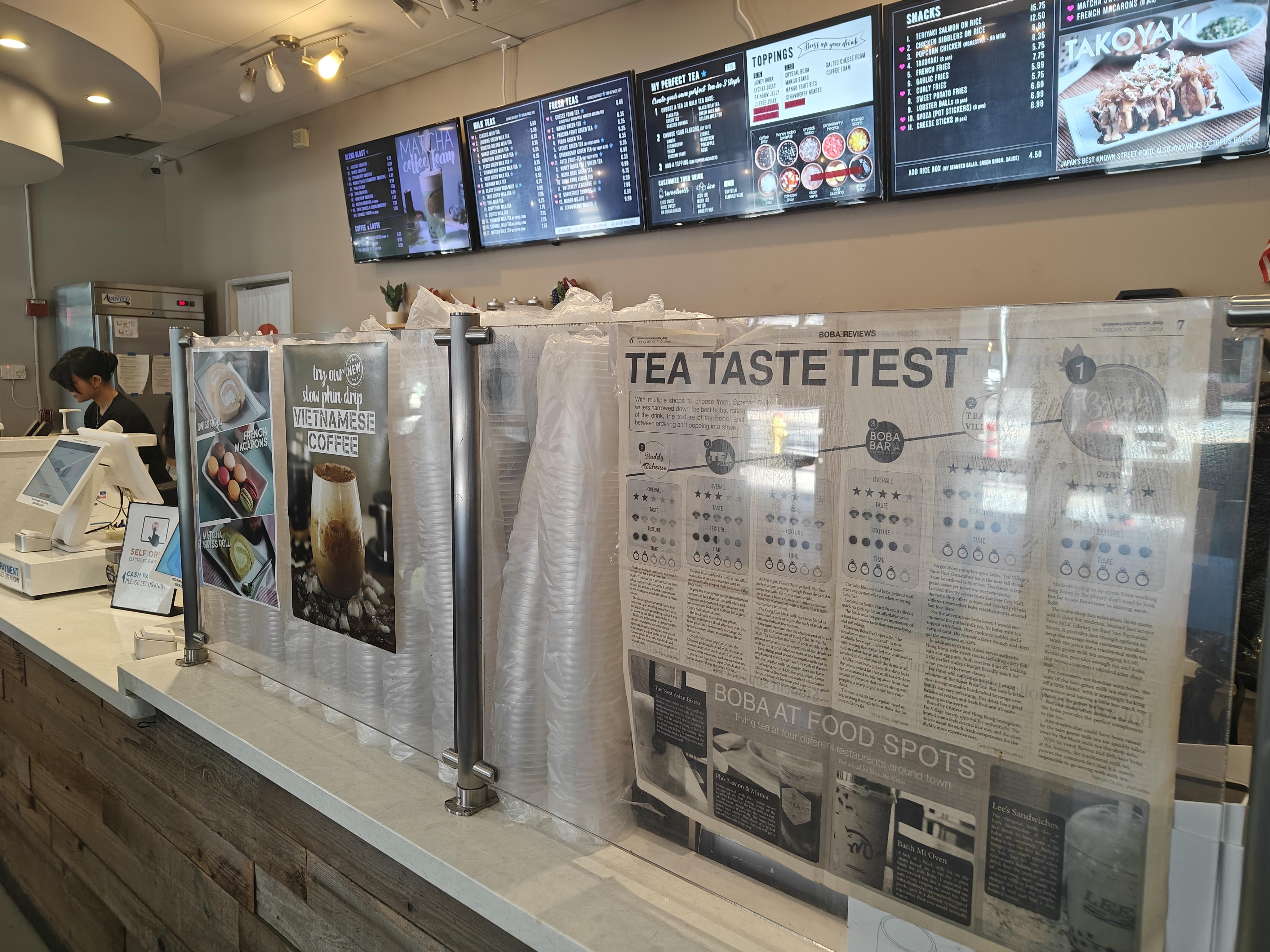 A printed newspaper with the headline TEA TASTE TEST and boba shop menu screens in the background