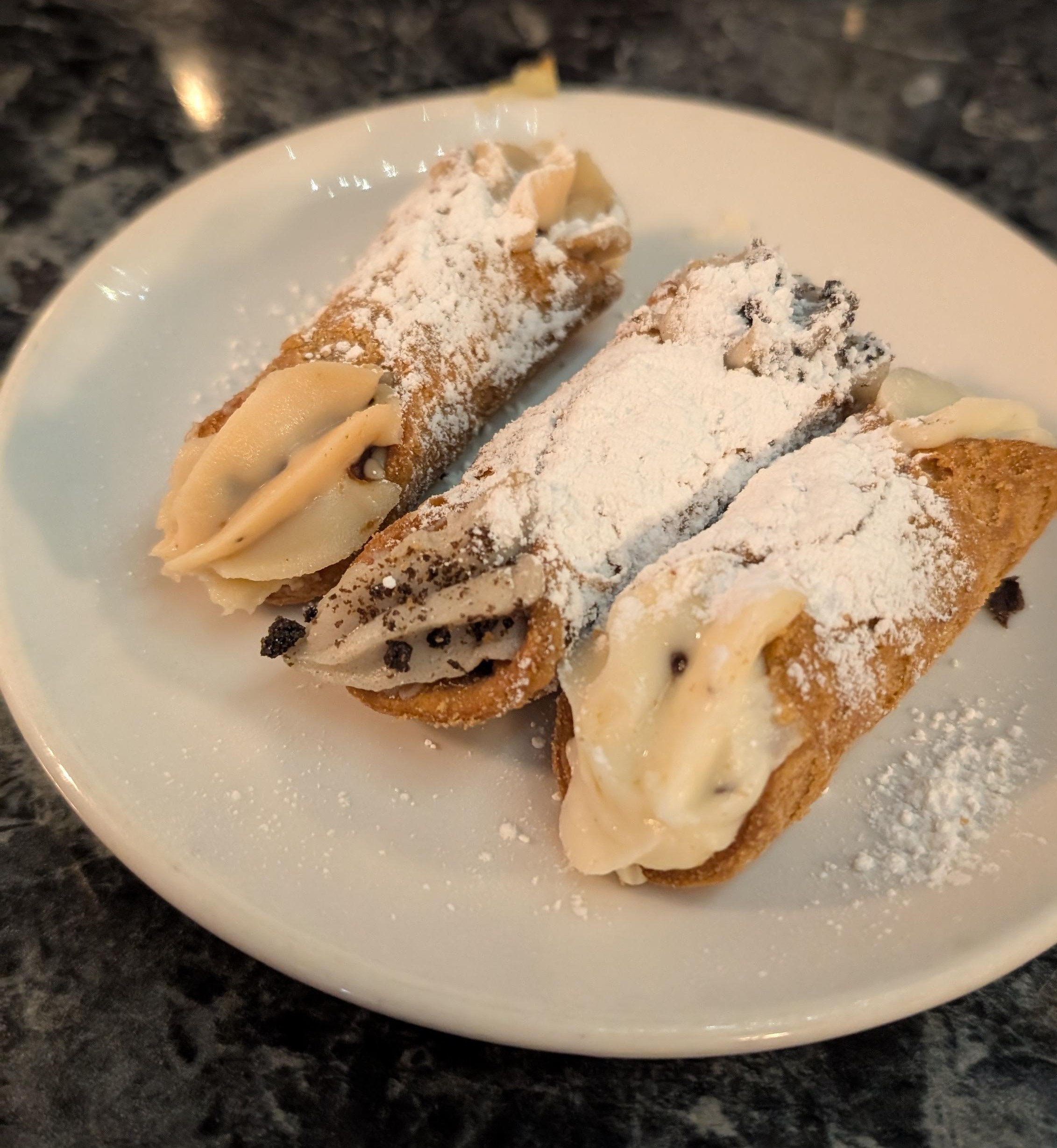 Three cannoli sitting on a plate. Flavors from left to right are hazelnut, cookies and cream, and original. All are covered in powdered sugar.