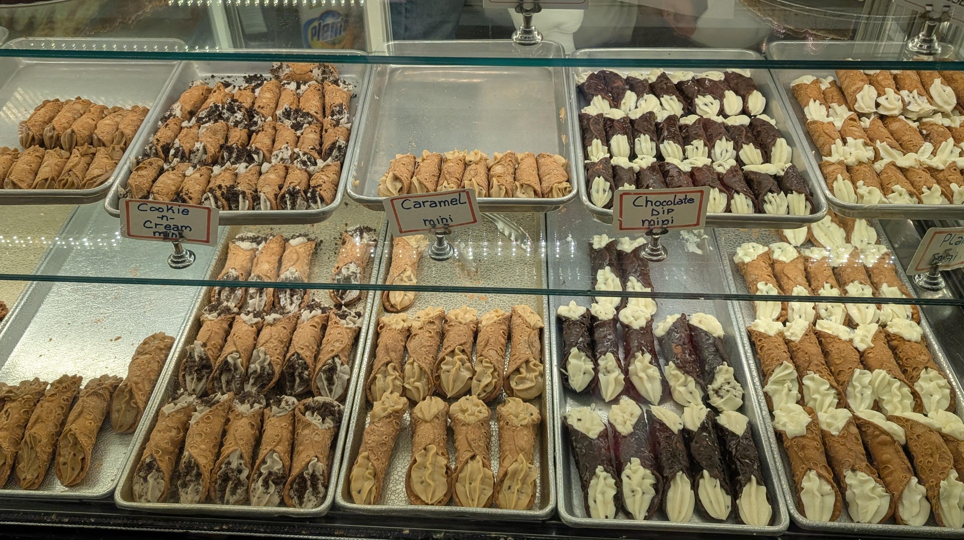 A bakery display case with trays of cannoli, with signs for cookie and cream, caramel and chocolate dip.