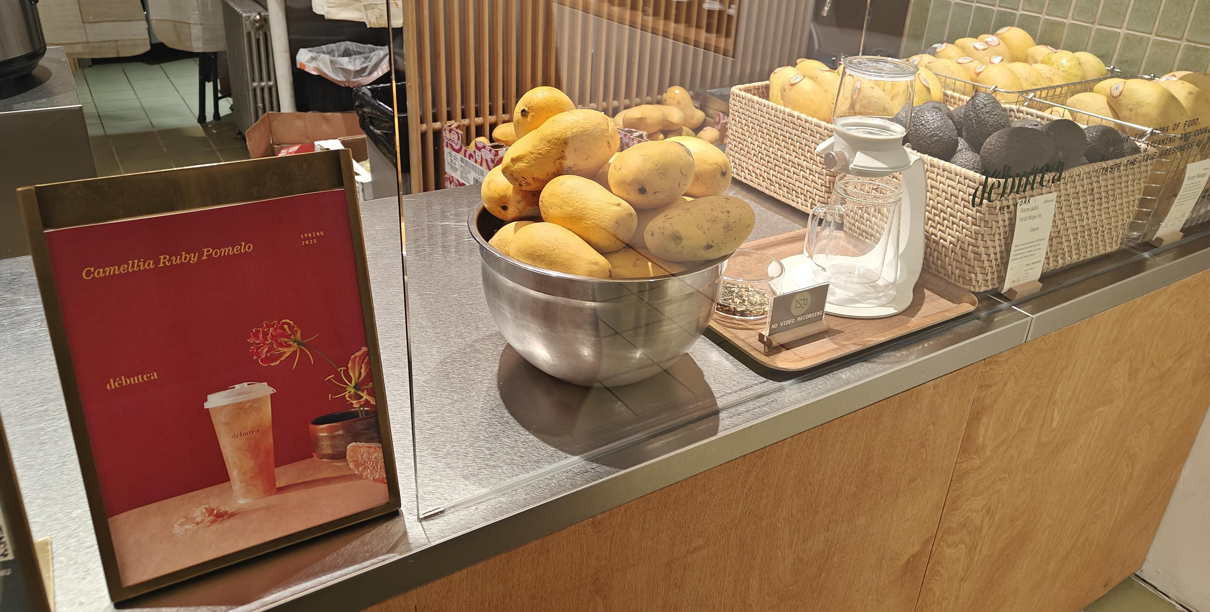 Boba shop counter, on the left there's a sign advertising the camellia ruby pomelo tea for spring 2025 and on the right are baskets of mangoes and avacados.