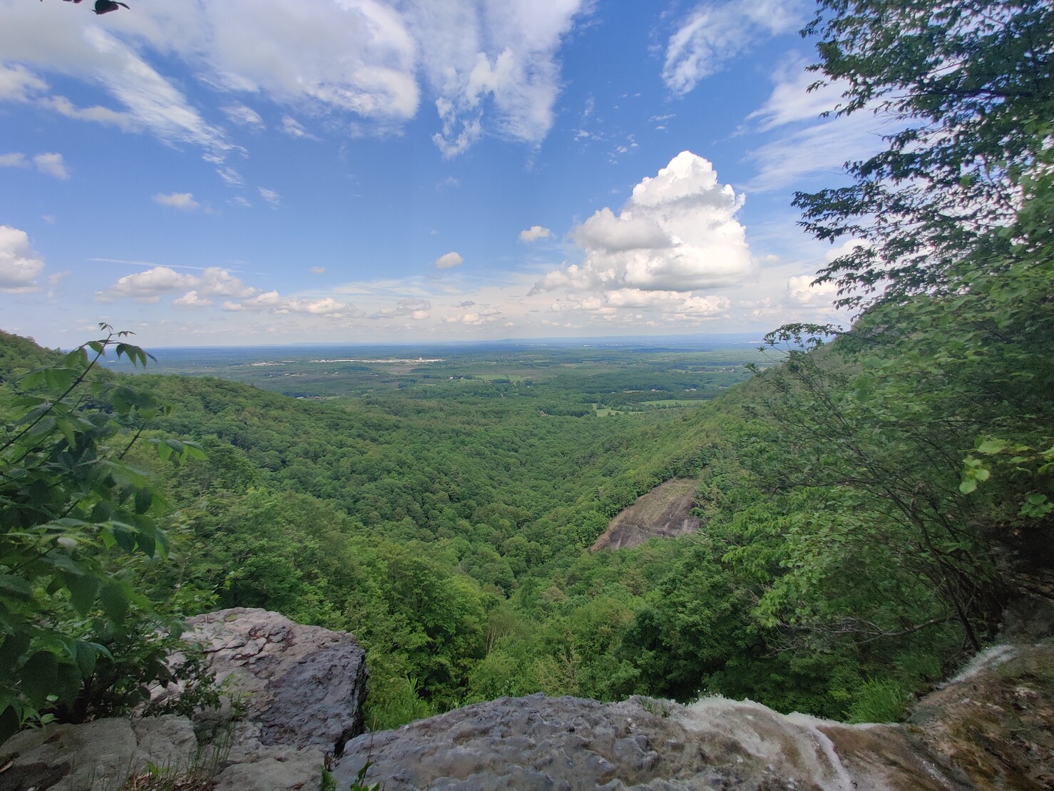 Sunny skies with light clouds above a lush green ground taken from a cliff with a waterfall