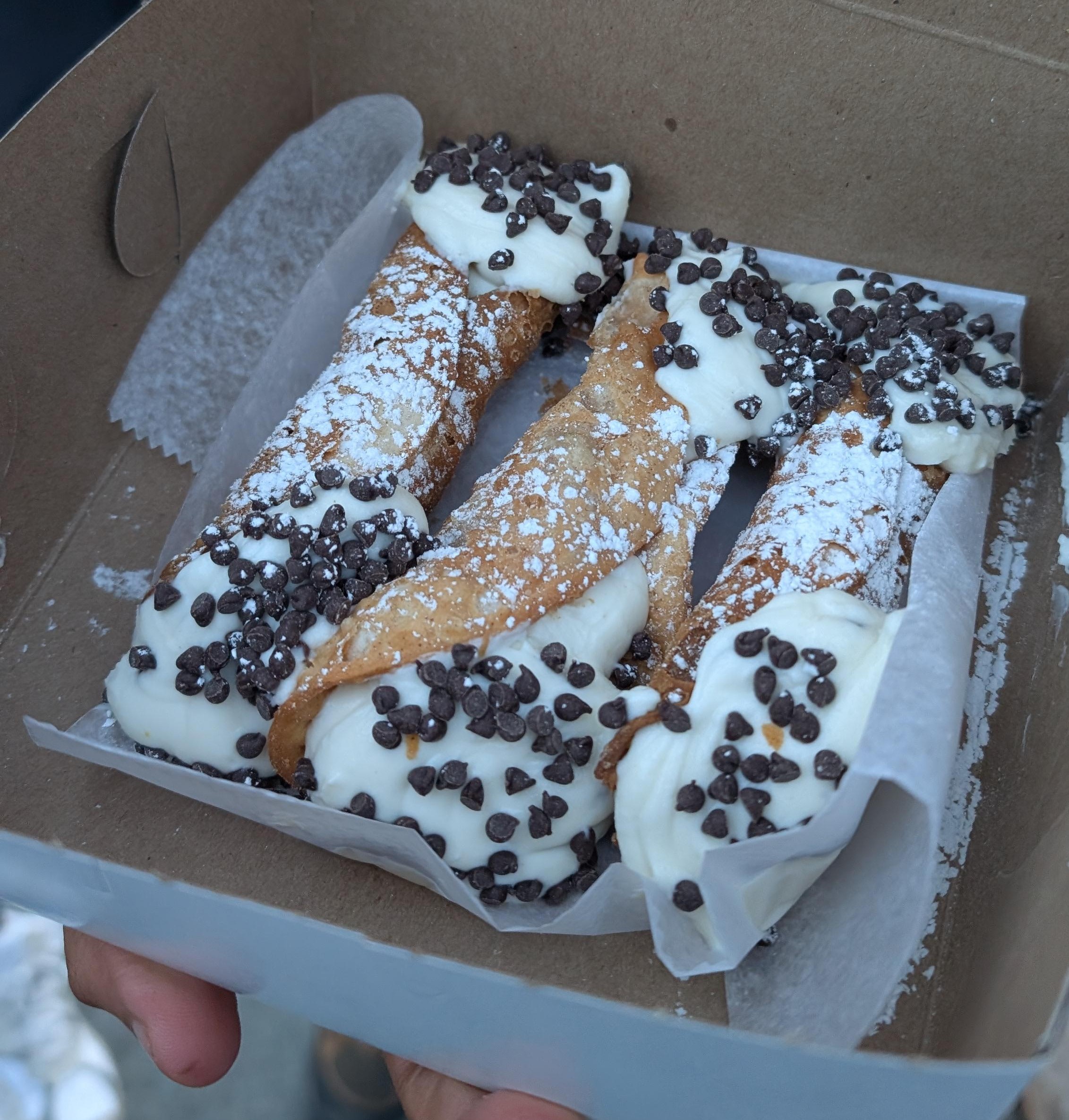 Three cannoli side-by-side in an open box. All three have mini chocolate chips on the ends and are covered in powdered sugar